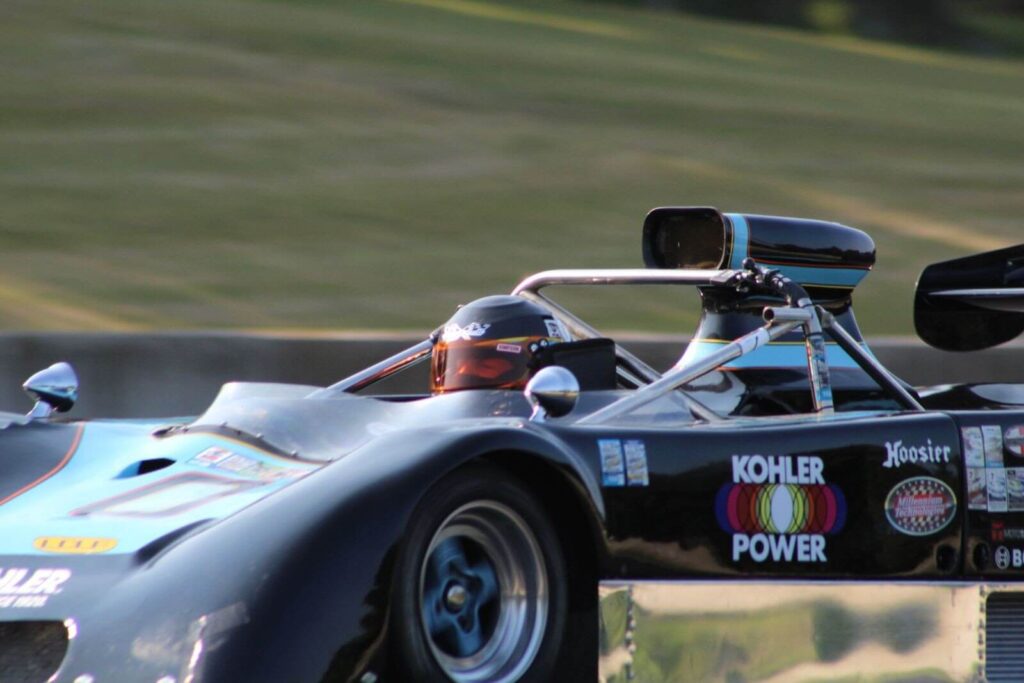 A black race car with blue and orange accents speeds along a track, its driver wearing a helmet with a vibrant design. The car features logos like "Kohler Power" and "Hoosier" on its side, with a low, aerodynamic body and spinning wheels capturing the motion.